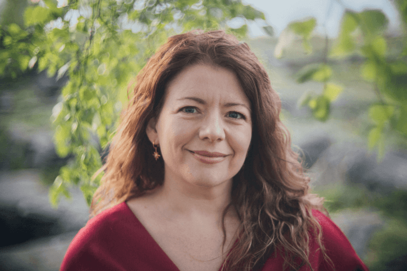 Moira Fuller, Business Mentoring expert, smiling outdoors in red top with green leaves in background