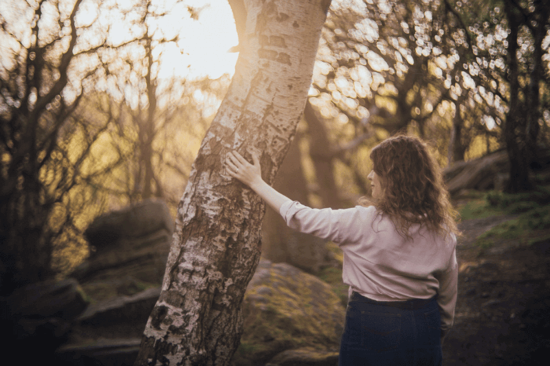 Moira Fuller, Business Mentoring coach, standing in woodland at sunset with hand on tree