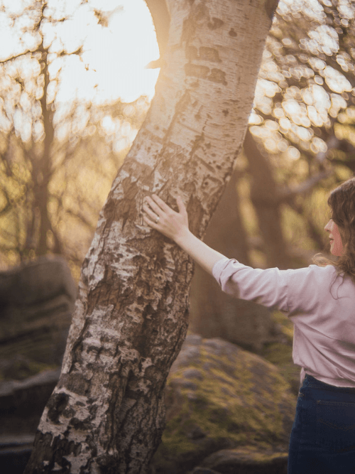 Moira Fuller, Business Strategist in the UK, standing in woodland with hand on tree at sunset